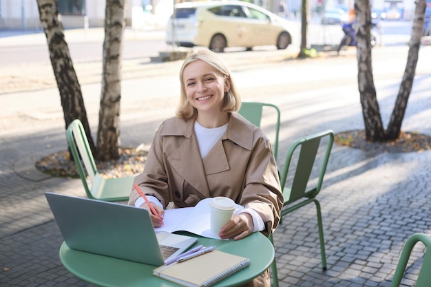Фото Портрет молодой женщины, работающей над проектом, сидящей на открытом воздухе в кафе, пьющей кофе с помощью ноутбука