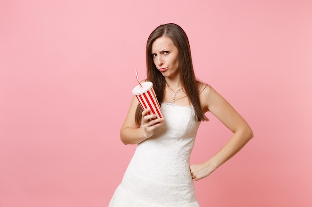 Portrait of offended irritated woman in white dress standing holding plastic cup with cola or soda