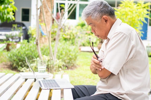 Portrait of old elderly Asian man using a computer laptop in the backyard for learning new skill after retired. Concept of no Ageism and not be late for learning.