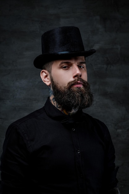 Portrait of an old-fashioned bearded man with tattoos on his neck wearing a top hat, isolated on a dark background.