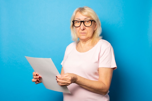 Portrait of an old friendly woman in casual t-shirt and glasses holding a sheet of paper in her hands on an isolated blue wall. emotional face. concept letter, notice