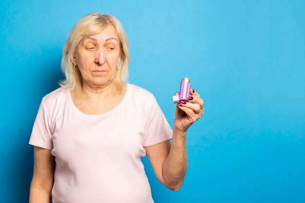 Portrait of an old friendly woman in a casual t-shirt holds an inhaler and looks at it on an isolated light wall. Emotional face. Asthma, allergy concept