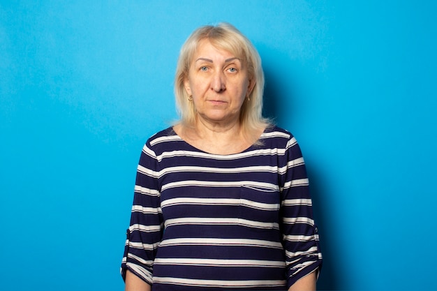 Portrait of an old friendly woman with a serious face in a casual t-shirt on an isolated blue wall. Emotional face