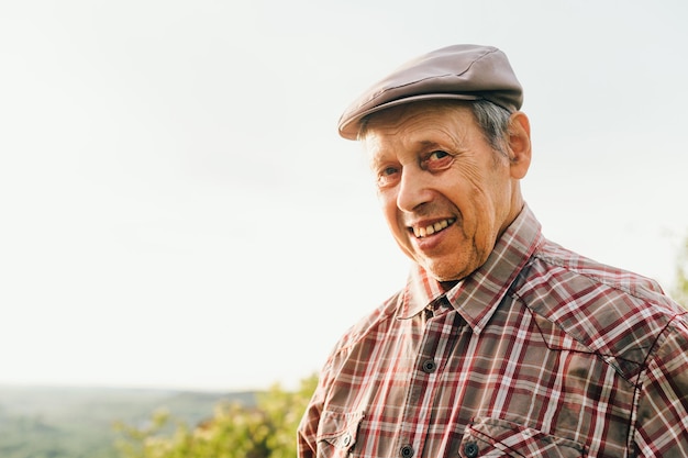 Portrait of old happy man in cap and shirt outdoors at sunset\
looking at camera and smiling retired man posing at camera close up\
photo portrait of an old positive grandfather