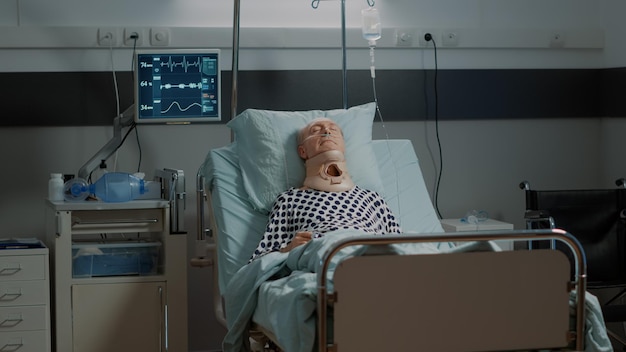 Portrait of old man with cervical neck collar resting in hospital ward bed at recovery facility. Senior patient with nasal oxygen tube and oximeter laying after surgery procedure