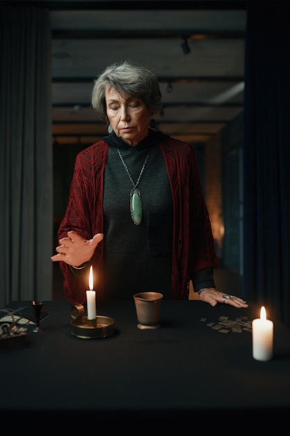 Portrait of old mature gypsy witch standing at table with psychic tools