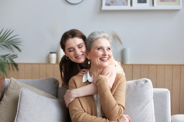 Portrait of old mother and mature daughter hugging at home. Happy senior mom and adult daughter embracing with love on sofa.