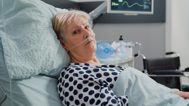 Portrait of old patient laying in bed with nasal oxygen tube\
and heart rate monitor for recovery treatment. senior woman with\
illness looking at camera and getting cure for healthcare