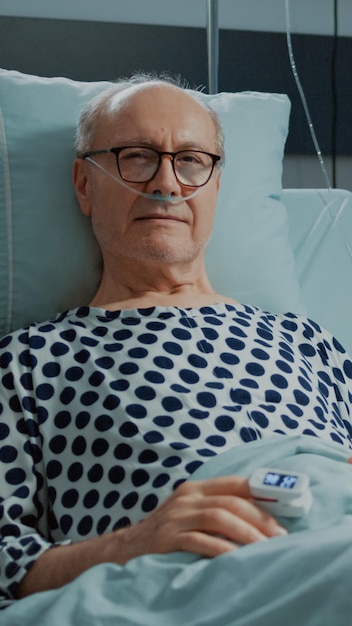 医学的健康問題のために鼻酸素チューブを備えたベッドで病棟に座っている病気の老人患者の肖像画。治療と相談のために医師と看護師を待っている男
