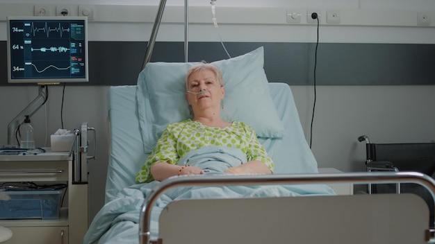 Portrait of old woman sitting in hospital ward bed to cure
disease, being hospitalized at intensive care. patient with nasal
drip bag and heart rate monitor for healthcare and medicine.