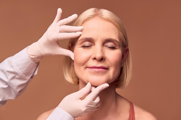 Photo portrait of an older adult woman with closed eyes on a beige background hands in white gloves and a medical gown show a face that has wrinklesface lift