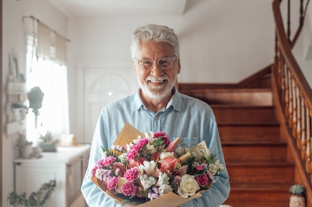 彼の妻やガールフレンドに与えるために花を持っている一人の幸せでかわいい老人の肖像画。バレンタインデーの贈り物やプレゼントを楽しんで家でカメラを見ているシニア