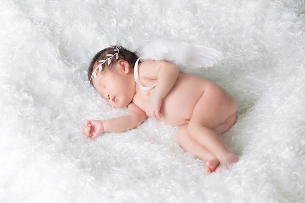 Portrait of a one month old sleeping, newborn baby girl. She is wearing a crown headband, feather angel wings, and sleeping on a white blanket. Concept portrait studio fashion newborn.