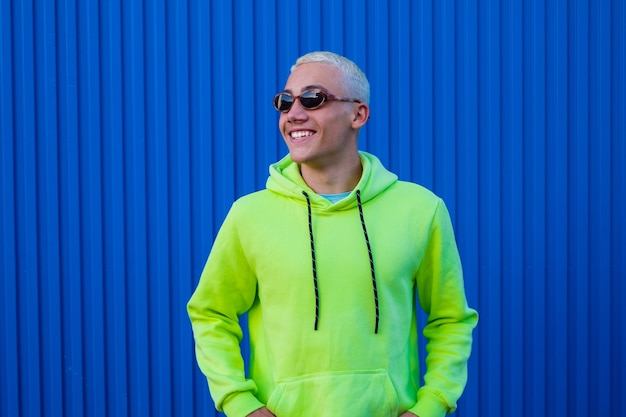 Portrait of one young man or teenager smiling and having fun wearing trendy sun glasses and lime sweater with blue colorful background - cheerful people millennial enjoying
