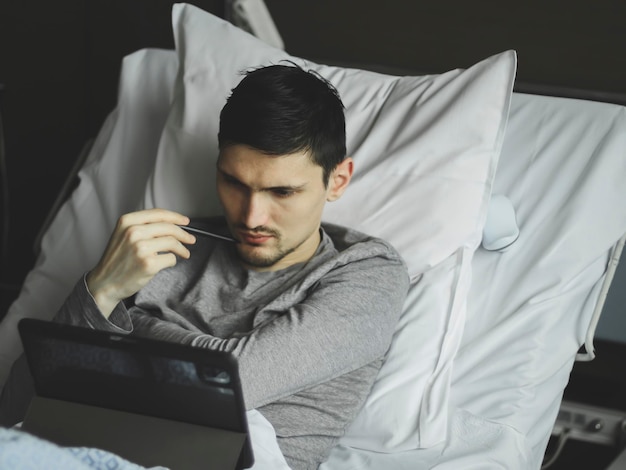 Portrait of one young pensive caucasian guy lying in bed with a tablet in a hospital room near the window top view closeup