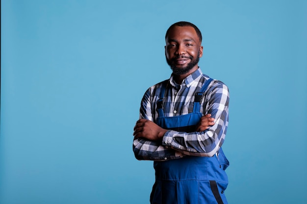 Портрет оптимистичного афроамериканского строителя, позирующего со скрещенными руками в студии. Профессиональный строитель в клетчатой рубашке и комбинезоне выглядит уверенно.