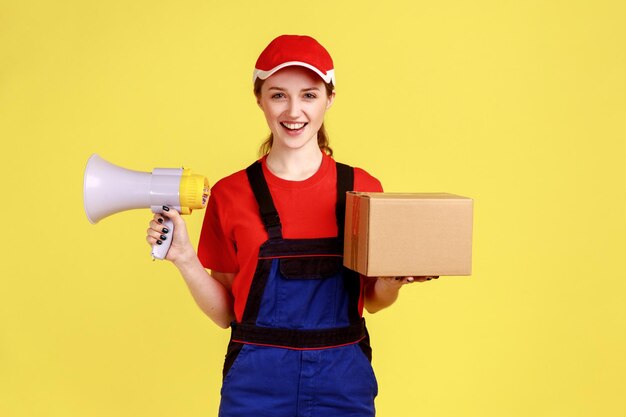 Portrait of optimistic courier woman standing holding cardboard box and megaphone looking at camera with happy expression wearing overalls Indoor studio shot isolated on yellow background