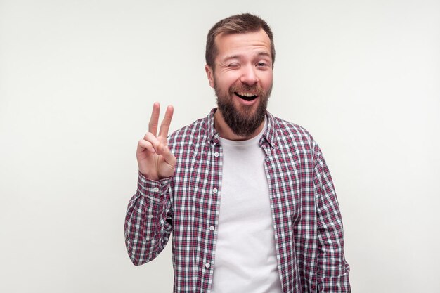 Portrait of optimistic funny bearded man in plaid shirt winking at camera smiling playfully and showing victory symbol with fingers number two or v sign studio shot isolated on white background