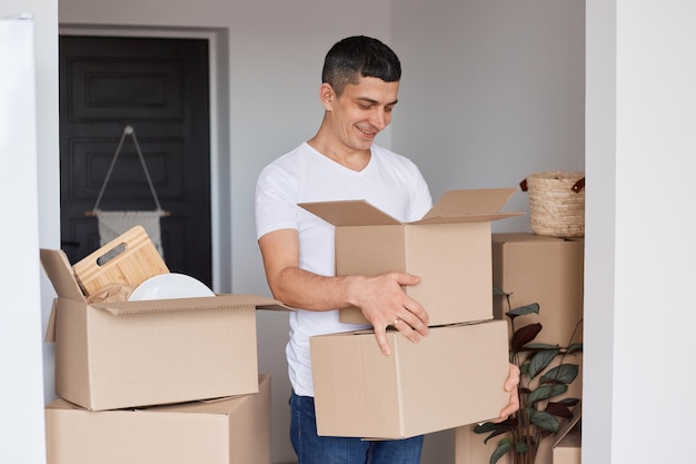 Portrait of optimistic handsome man wearing white T-shirt standing with cardboard boxes with personal stuff, looking inside with happy expression, relocation, real estate.