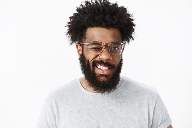 Portrait of optimistic and joyful handsome african american guy in good mood winking at front friendly and grinning making positive impression having good day over gray wall