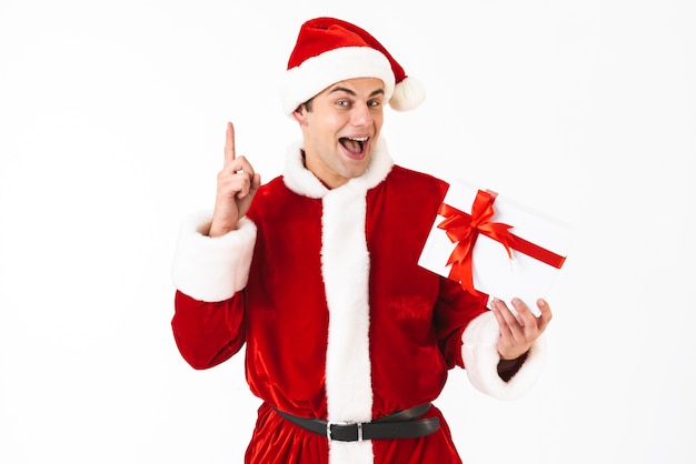 Portrait of optimistic man 30s in santa claus costume and red hat holding present box