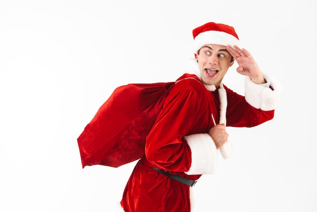 Portrait of optimistic man 30s in santa claus costume and red hat running with gift bag over shoulder