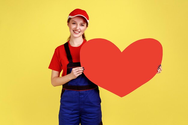 Portrait of optimistic worker woman standing holding big red heart in hands, looking at camera with happy expression, wearing overalls and red cap. Indoor studio shot isolated on yellow background.