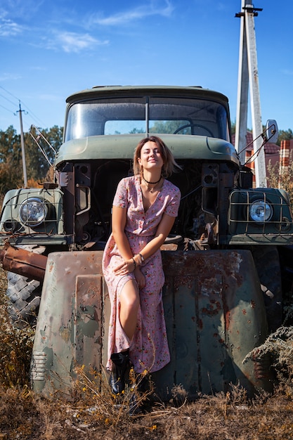 Portrait outdoor atmospheric lifestyle photo of young beautiful\
dark haired woman in a pink dress in a floral print against the\
backdrop of an old truck car