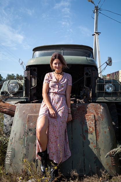 Portrait  outdoor atmospheric lifestyle photo of young beautiful  darkhaired woman in a pink dress in a floral print against the backdrop of an old truck car . Autumn walking concept