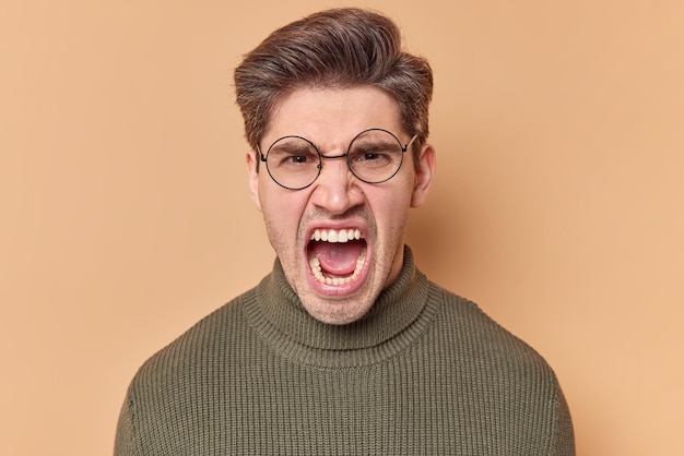 Portrait of outraged young European man screams angrily keeps mouth opened expresses negative emotions feels irritated wears round spectacles and sweater isolated over brown studio background