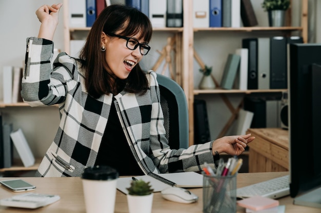 写真 机に座って喜んでいるアジアの女性マネージャーのポートレートは、オフィスのコンピューターでオンラインビジネスの成功を祝うために指を鳴らして踊っています。