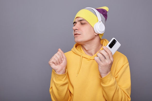 Portrait of overjoyed Caucasian guy singing song and holding smart phone in hands clenched fist enjoying song keeps eyes closed wearing wireless headset dancing isolated over grey background
