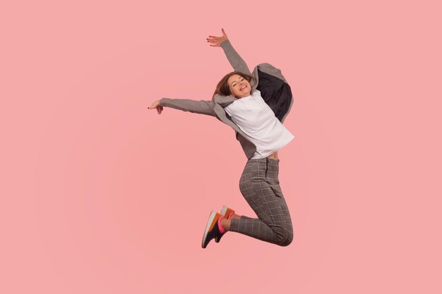 Portrait of overjoyed enthusiastic ecstatic businesswoman in elegant suit jumping carefree in air flying from happiness inspired by success freedom indoor studio shot isolated on pink background