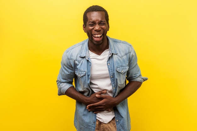 Portrait of overjoyed happy man in denim casual shirt with\
rolled up sleeves laughing hard and holding hands on stomach\
feeling amused by joke indoor studio shot isolated on yellow\
background