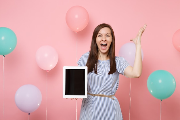Portrait of overjoyed happy woman in blue dress holding tablet pc computer with blank empty screen spreading hands on pastel pink background with colorful air balloons. Birthday holiday party concept.