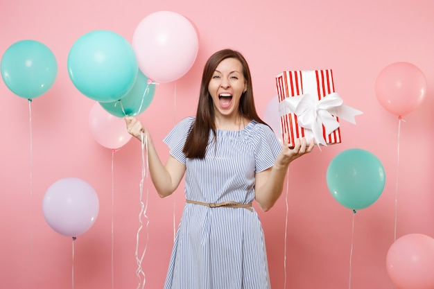 Portrait of overjoyed happy woman wearing blue dress holding red box with gift present and colorful air balloons on bright pink background. Birthday holiday party, people sincere emotions concept.