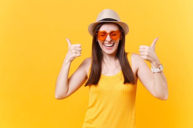 Portrait of overjoyed laughing young woman in straw summer hat, orange glasses showing thumbs up, copy space isolated on yellow background. People sincere emotions, lifestyle concept. Advertising area