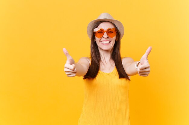 Portrait of overjoyed laughing young woman in straw summer hat, orange glasses showing thumbs up, copy space isolated on yellow background. People sincere emotions, lifestyle concept. Advertising area