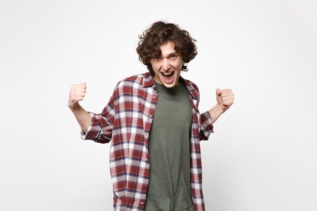 Portrait of overjoyed screaming young man in casual clothes clenching fists like winner isolated on white wall background in studio. People sincere emotions, lifestyle concept. Mock up copy space.