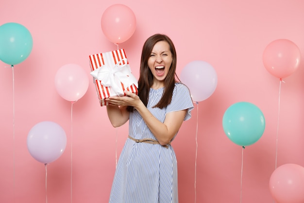 Portrait of overjoyed young woman in blue dress screaming holding red box with gift present on pastel pink background with colorful air balloon. Birthday holiday party, people sincere emotion concept.