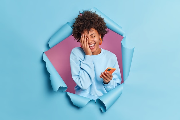 Portrait of overjoyed young woman makes face palm smiles broadly keeps eyes closed uses smartphone for texting expresses positive authentic emotions breaks through blue paper wall