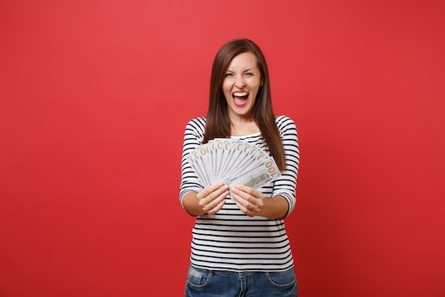 Portrait of overjoyed young woman in striped clothes screaming holding bundle lots of dollars cash money isolated on red wall background. People sincere emotions lifestyle concept. Mock up copy space.