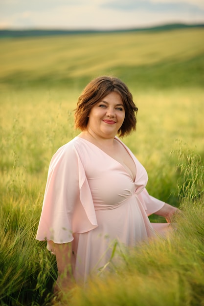 Портрет полной женщины в розовом платье. Женщина стоит в зеленом пшеничном поле
