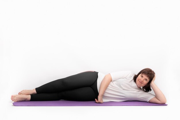 Portrait of an overweight young woman lies and looks at the camera on yoga mat on white background. Plus size model goes in for sports.