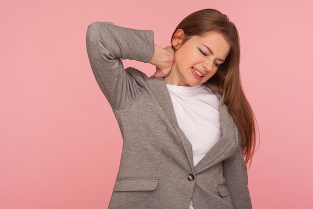 Portrait of overworked stressed young woman in business suit\
touching painful neck suffering from aching muscles hurting\
shoulder feeling unhealthy indoor studio shot isolated on pink\
background