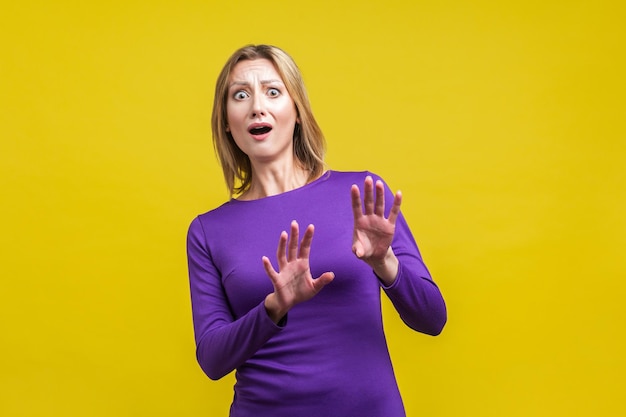 Photo portrait of panicked scared woman in tight purple dress raising hands looking at camera with horrified eyes and open mouth sudden fright indoor studio shot isolated on yellow background