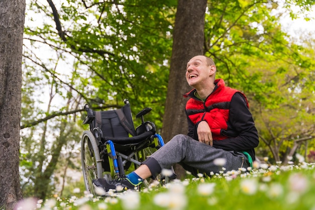 Portrait of a paralyzed young man sitting on the grass next to the wheelchair in a daisy flower smiling and enjoying nature