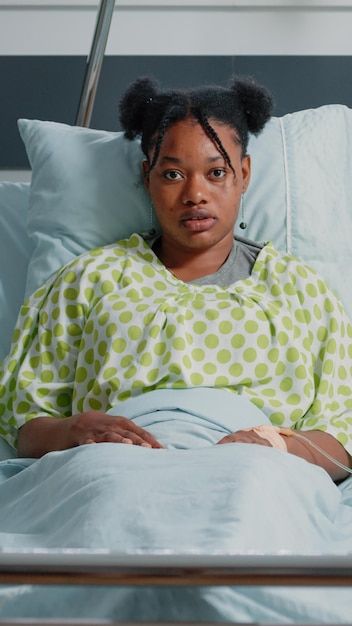 Portrait of patient with disease receiving medical treatment in hospital ward bed. Young woman with heart rate monitor and IV drip bag looking at camera while healing from sickness