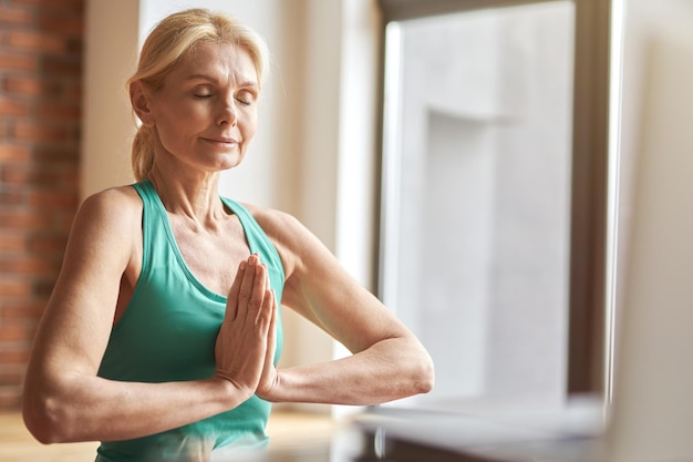 Ritratto di una donna bionda matura pacifica che pratica yoga meditando con gli occhi chiusi a casa nel