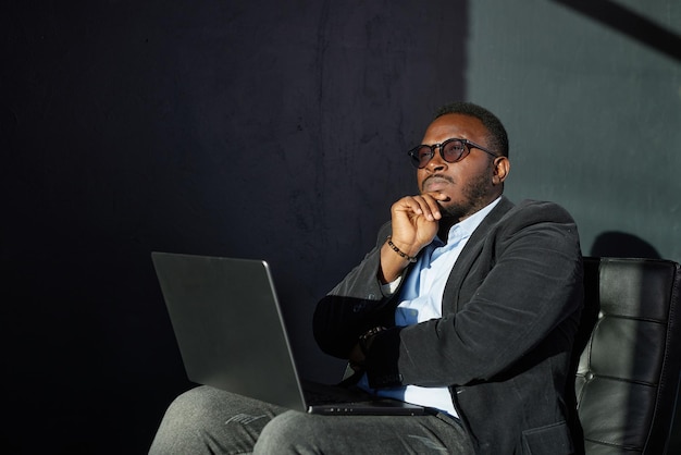 Портрет задумчивого африканского бизнесмена в очках, сидящего за столом с ноутбуком в домашнем офисе